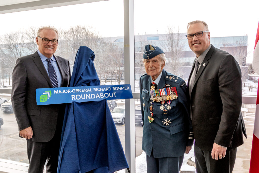 Three people, one of whom is in a military uniform, stand next to a sign reading: Major-General Richard Rohmer Roundabout.