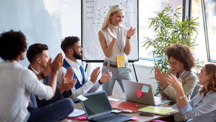 A person standing next to a board room table as they and those sitting around the table smile and clap their hands