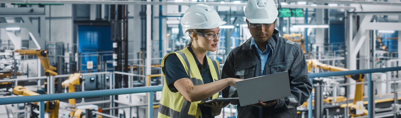 Manufacturing leadership team wearing hard hats and safety goggles holding a laptop and tablet while standing in front of a railing overlooking the machinery and equipment
