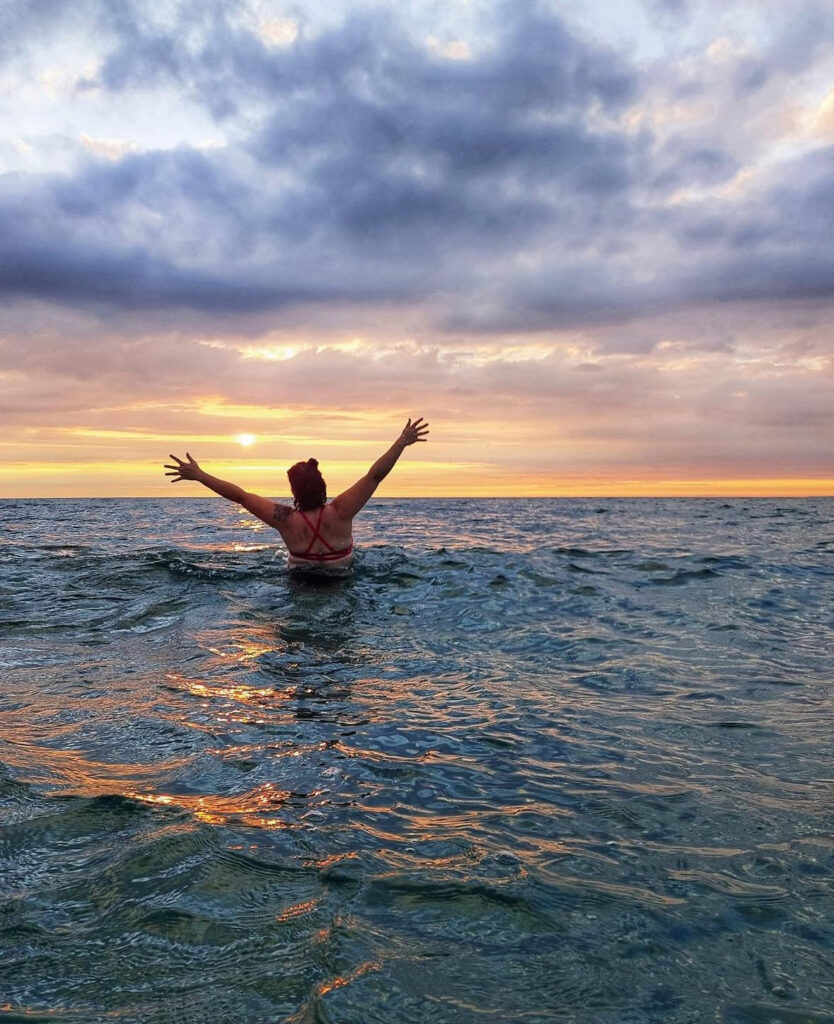 A person stands in a lake up to their waist, facing away from the camera, and puts their arms in the air.