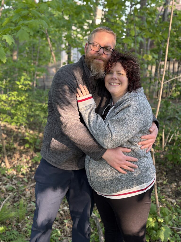 Two people stand together in a wooded area.