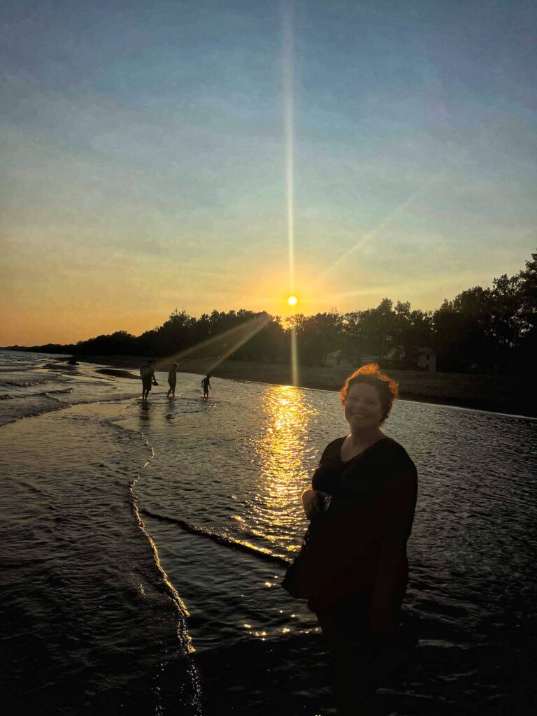 A person stands in a lake's shallow water and smiles with a sunset behind them in the distance.