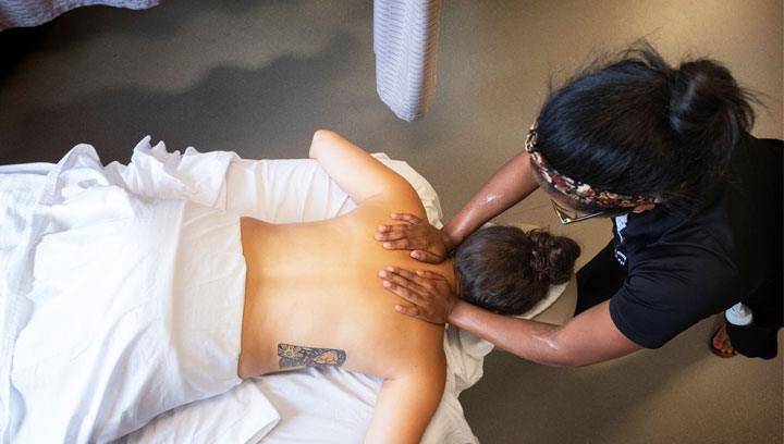 Massage Therapy student in a black collared shirt using her hands to massage the upper back of a laying down patient