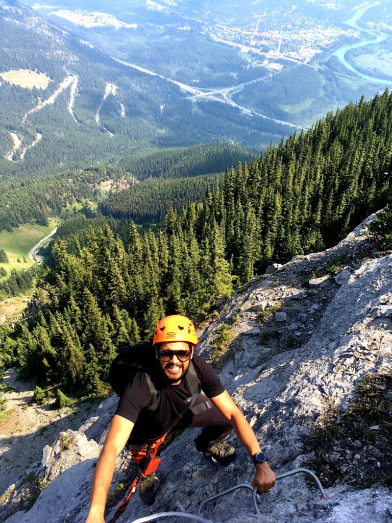 A person wearing all black, sunglasses and an orange helmet climbs up a mountain in a forested area.