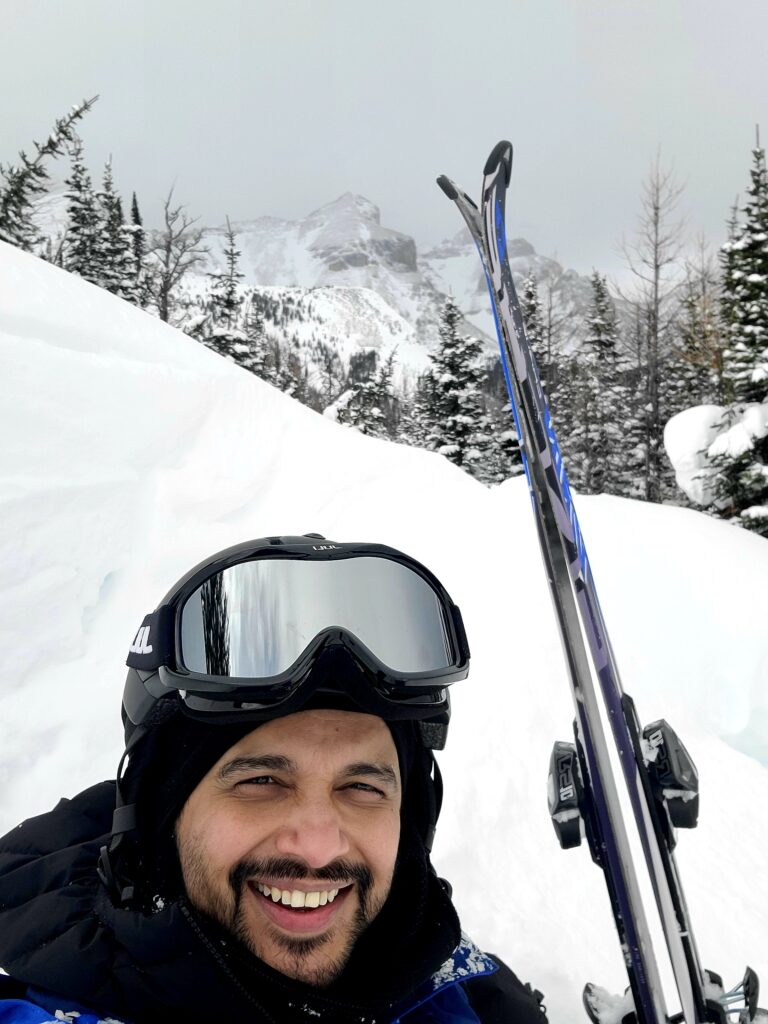 A selfie of a person wearing a winter coat and ski goggles and holding skis while standing outside on a snowy mountain.