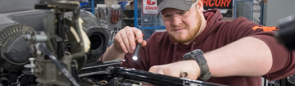 A Small Engine Mechanic wearing safety glasses while holding a flashlight and inspecting an engine in a work shop