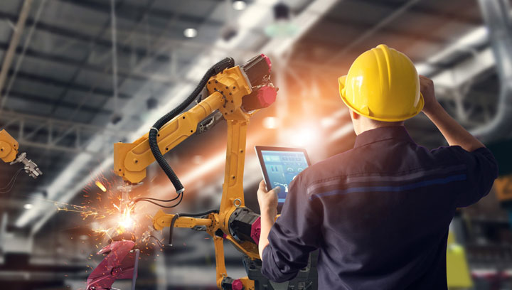 A Mechatronics specialist wearing a yellow construction hard hat holding a tablet and programming a robot