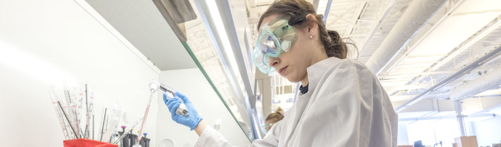 Medical lab tech wearing a white lab coat, blue latex gloves and green safety goggles pouring a solution into a test tube in a laboratory