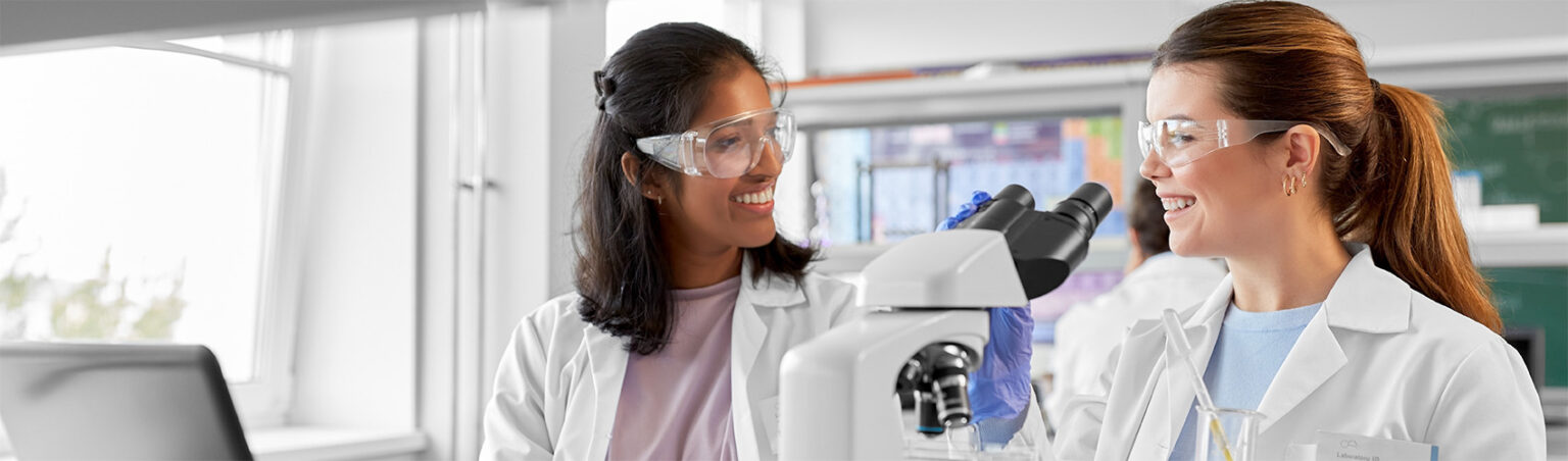 Two medical lab technicians in white collared lab coats and plastic safety goggles preparing to analyze a blood sample through a medical telescope