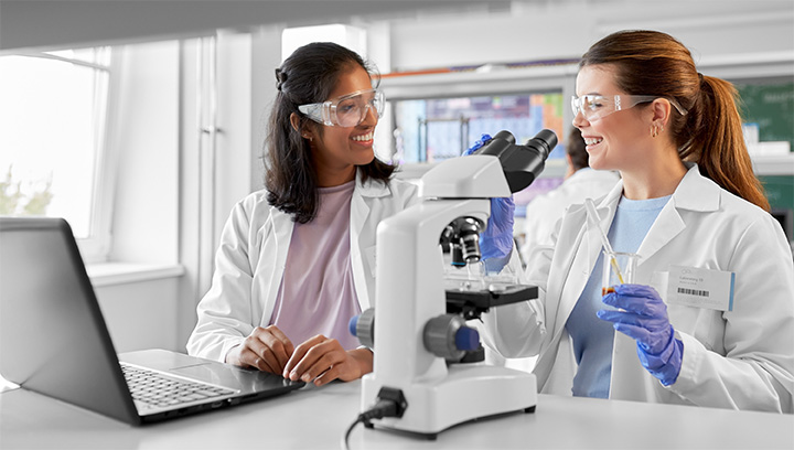 Two medical lab technicians in white collared lab coats and plastic safety goggles preparing to analyze a blood sample through a medical telescope