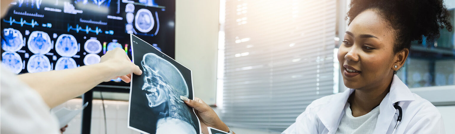 Person in a clinical setting reviews a head and neck X-ray film while another person points to an area of interest.