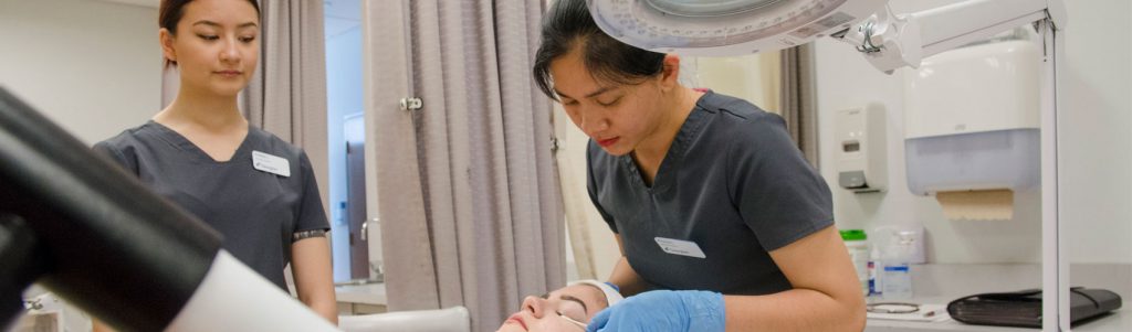 Two skin care specialists wearing gray scrubs and blue latex gloves applying a treatment to a patient's eyelids in a spa clinic