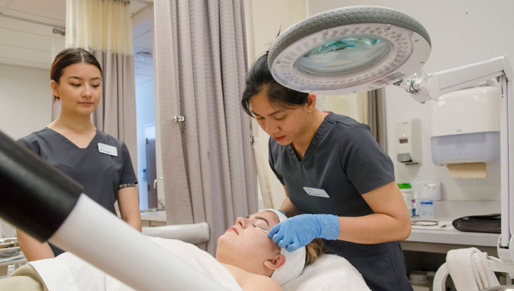 Two skin care specialists wearing gray scrubs and blue latex gloves applying a treatment to a patient's eyelids in a spa clinic