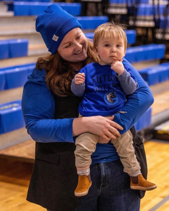 A person holds a toddler and smiles at them while standing in a gymnasium.