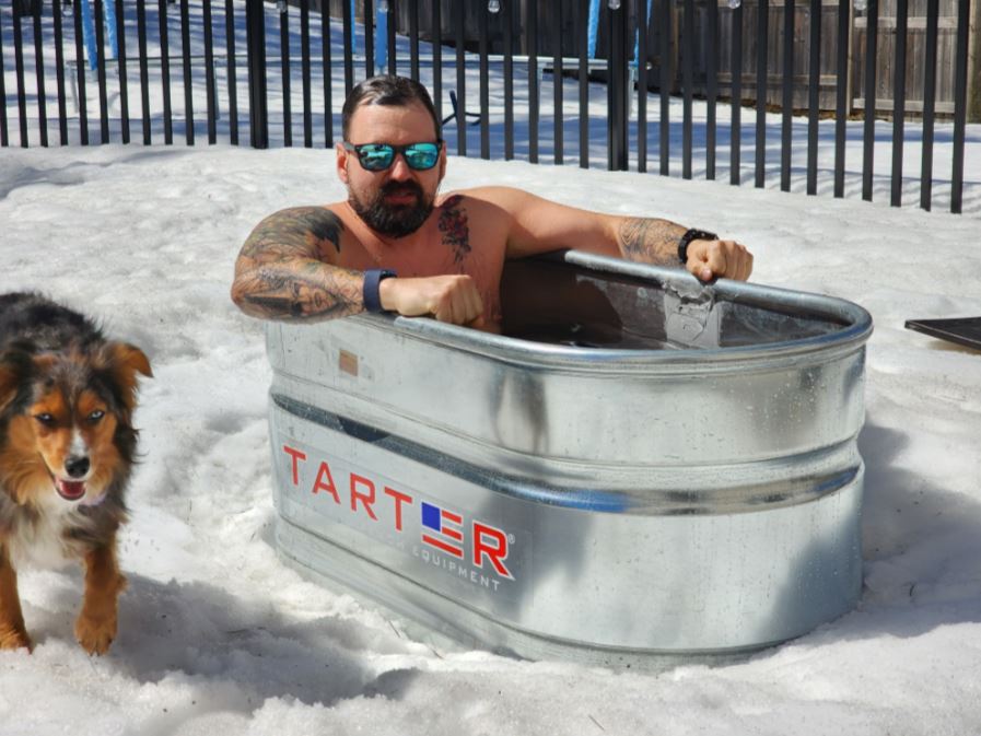 A person sits in an ice bath in a metal tub sitting outside in the snow, as a dog walks by.