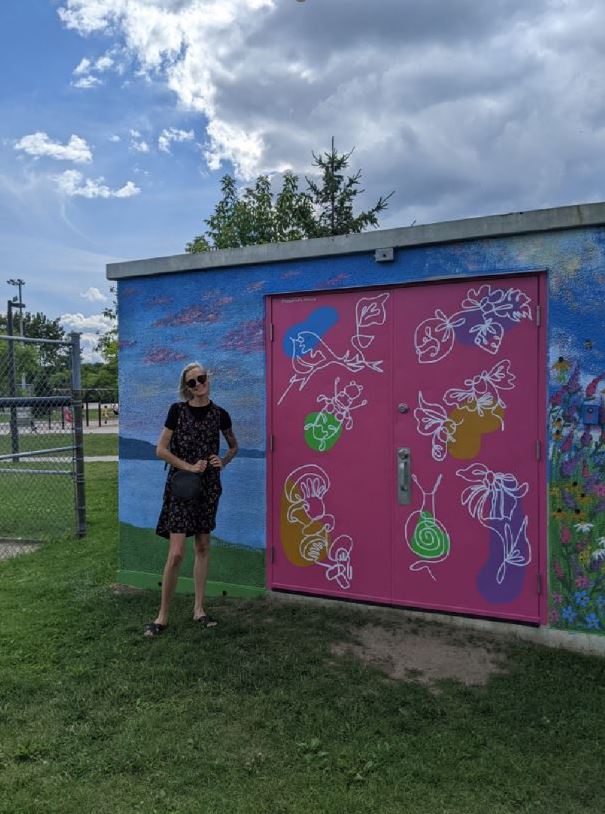 A person stands outside next to a small building with colourful artwork on it.