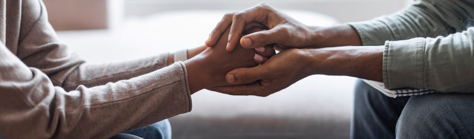 A grief counsellor sitting across from a client with their hands wrapped around the client's hands, offering support with the loss of a loved one