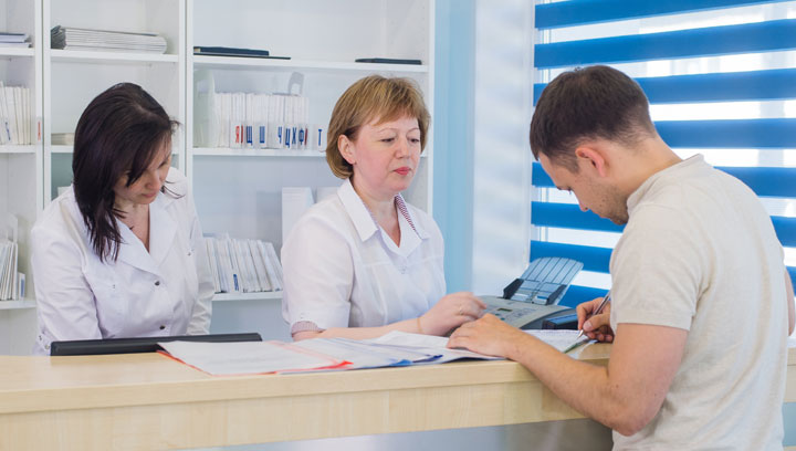 Two administrative professionals in a health-care setting wearing lab coats assisting a client over the counter with paperwork