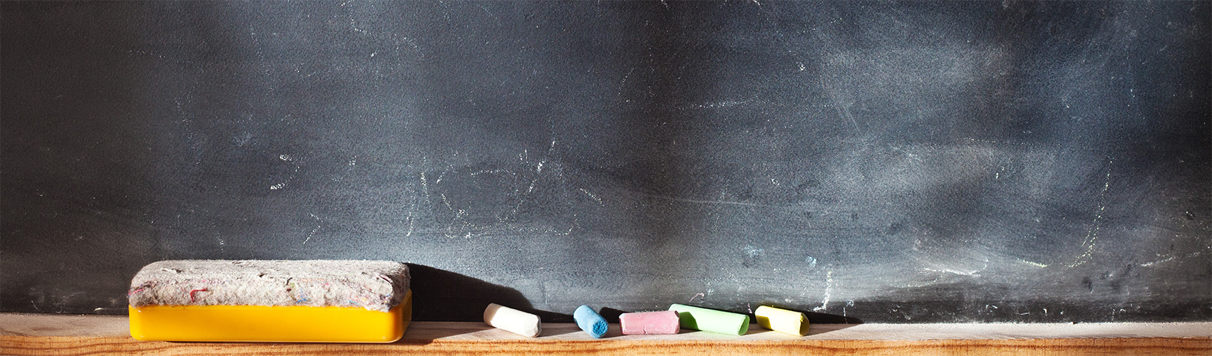 A black chalkboard with a wooden ledge holding an eraser and white, blue, red, green and yellow chalk, and chalk particles