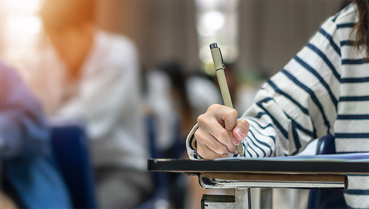 A group of students sitting at individual desks in a classroom setting writing with pens on a stapled package of paper