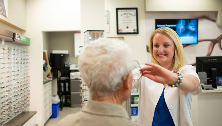 Optician with blonde hair and a white lab coat smiling while placing a pair of prescription eye glasses on a patient