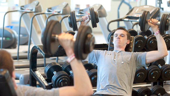A young man in a grey t-shirt lifting weights inside the Athletics and Fitness Centre at the СŶƵ Orillia Campus