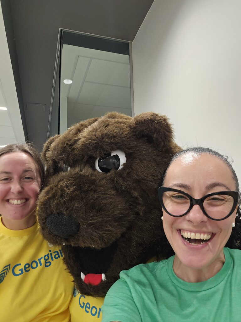 Two people take a selfie with a grizzly bear mascot.