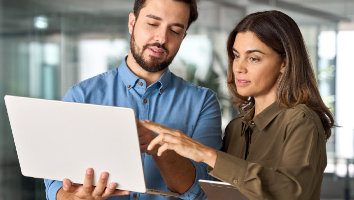 Two people in an office review information together on a laptop, with one person pointing at the screen during a discussion.