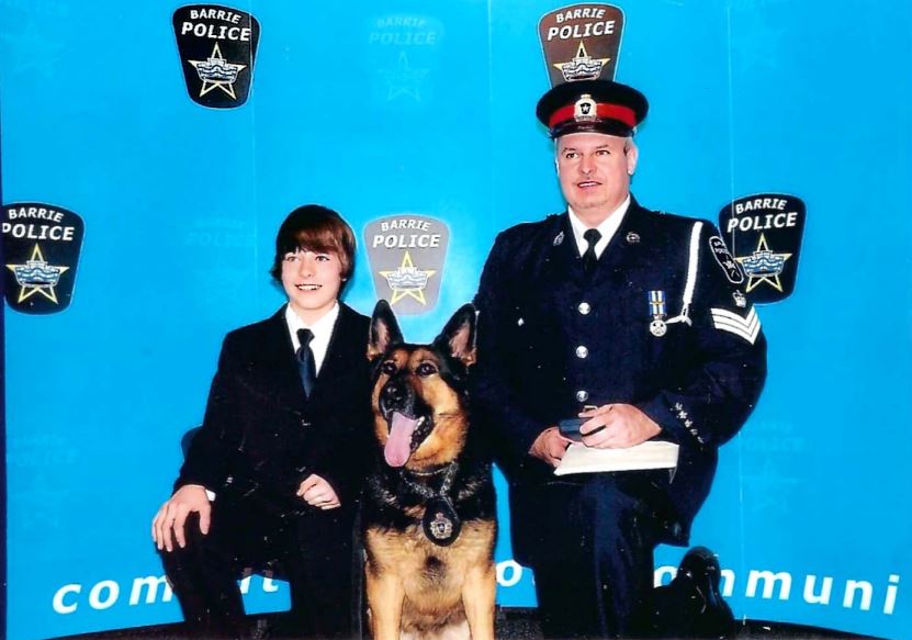 A child in a suit and an adult wearing a police officer's uniform kneel next to a German shepherd dog in front of a backdrop that reads "Barrie Police."