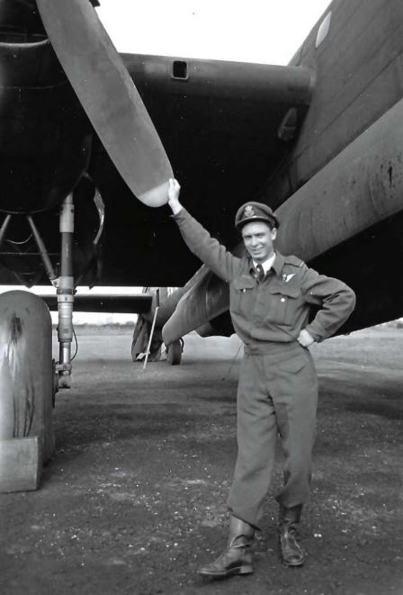 A black-and-white photo of a person in an air force uniform standing next to a large airplane with an arm reaching up to touch a propellor. 