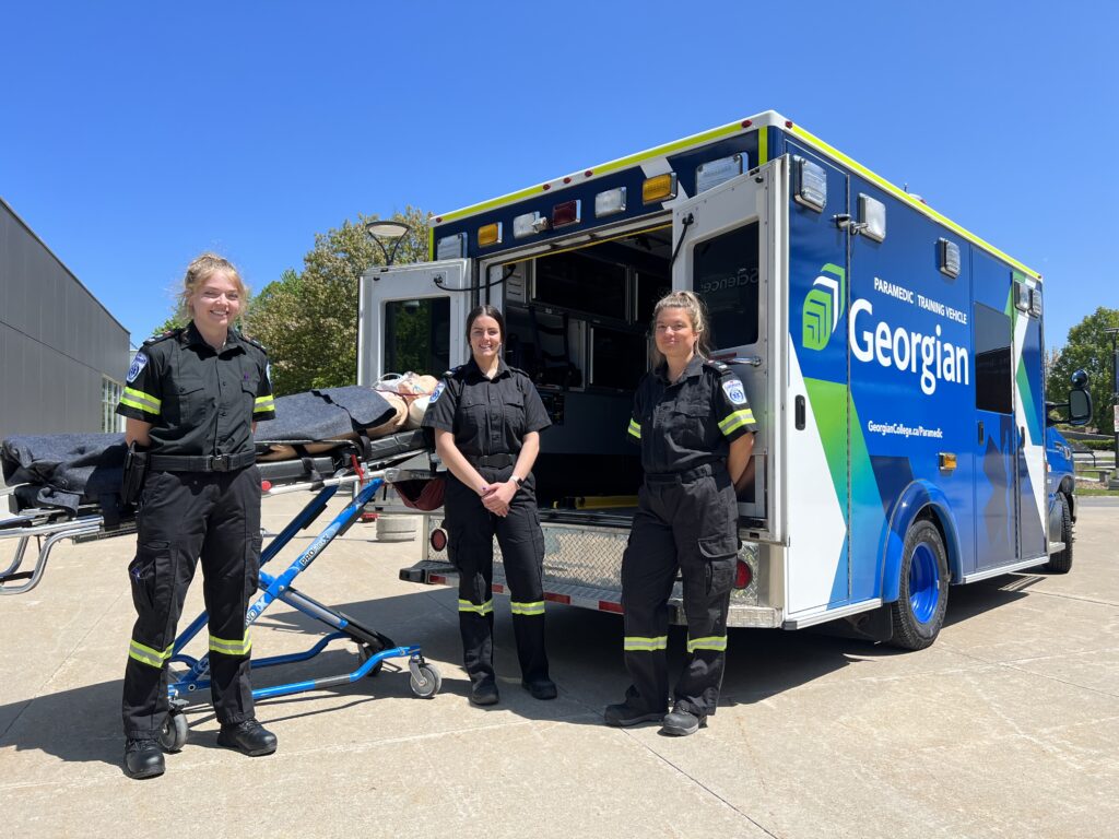 Three Paramedic students stand in front of a new training vehicle outside at Georgian College's Barrie Campus