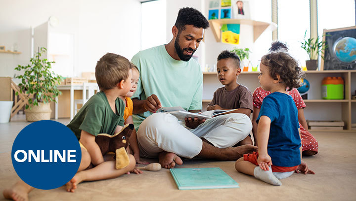 A person with dark hair and a beard sitting cross-legged on a floor in a daycare/classroom setting reading a book to a group of young children seated around