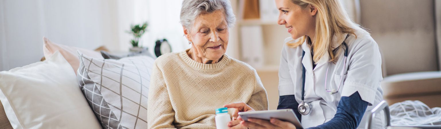 Personal Support Worker in scrubs holding a tablet while relaying medication instructions to an elderly patient in a care home