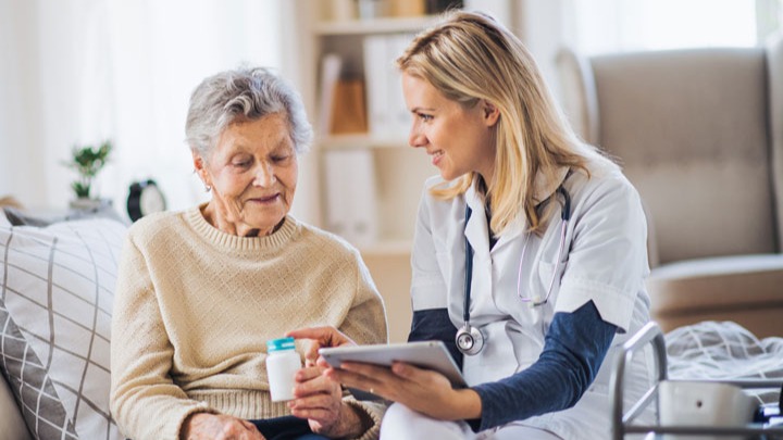 A young female presenting personal support worker with shoulder length straight blonde hair wearing scrubs is reading an older female presenting senior.