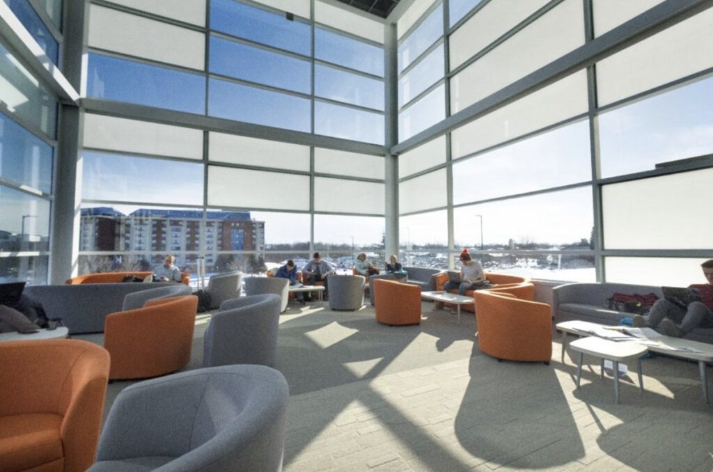 People sit on orange and grey chairs in a room with floor-to-ceiling windows. 