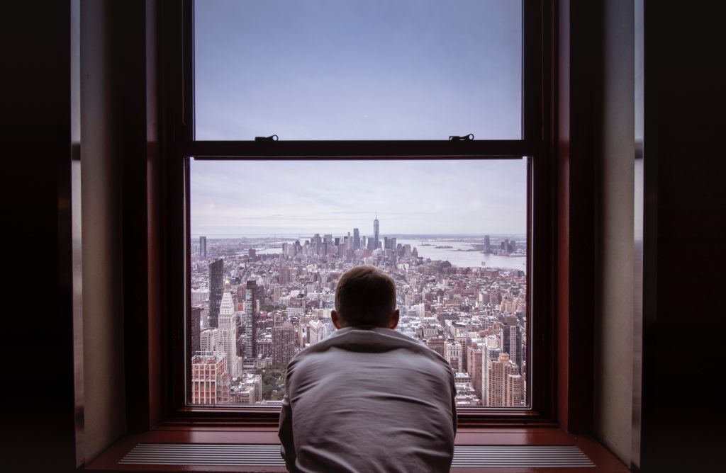 A man with his back turned, looking out a window overlooking a city landscape. 