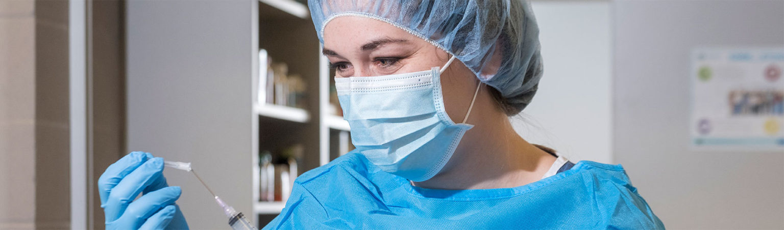 СŶƵ Pharmacy Technician student wearing scrubs, a face covering and hair mask preparing a vaccination syringe