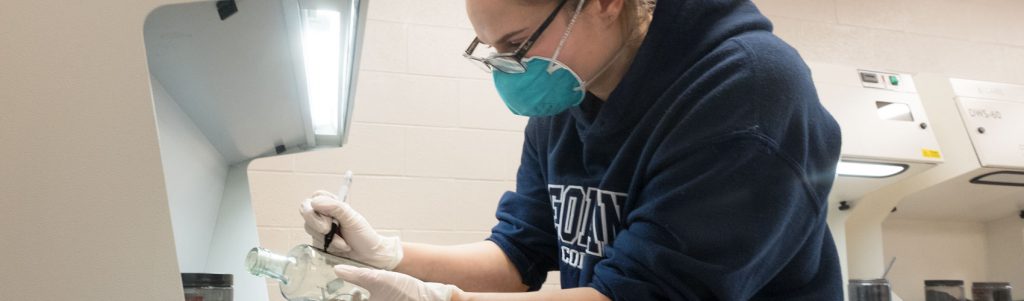 Police Foundations student wearing a navy Georgian hoodie, glasses and a mask labelling a glass bottle with a marker in a lab