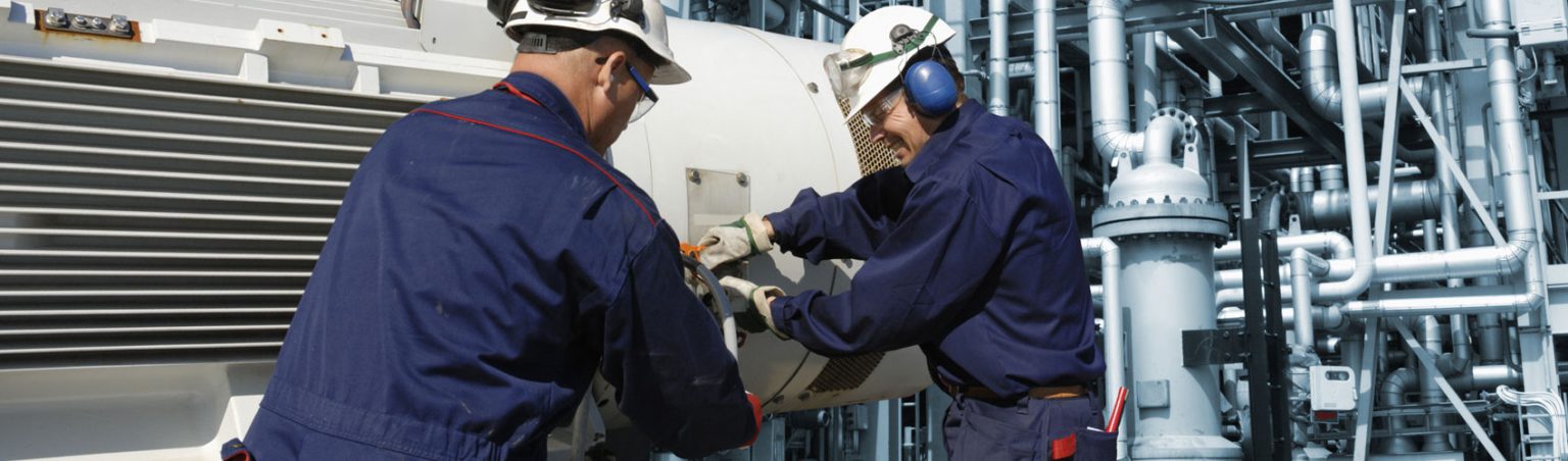 Two power engineering professionals wearing work jumpsuits and hard hats working on a piece of equipment