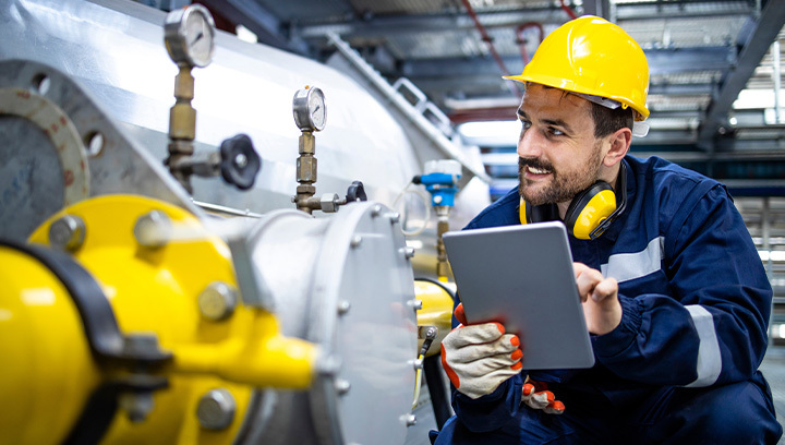 industrial worker wearing a yellow hard hat and protective headphones around his neck, inspecting machinery with pressure gauges inside a facility.