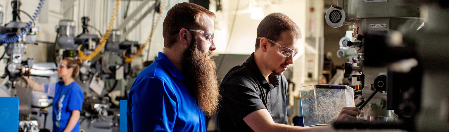 Two people wearing safety glasses working with industrial machinery in a workshop, focusing on precise technical tasks.