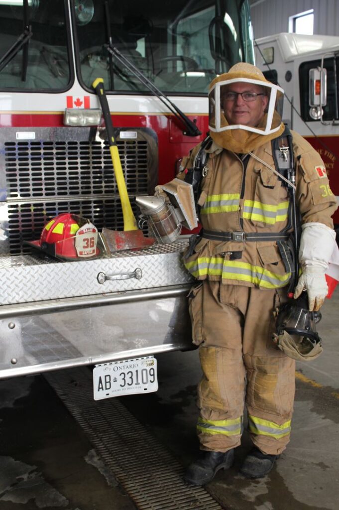 A person wearing a firefighter uniform and beekeeper's hat stands in front of a fire truck.