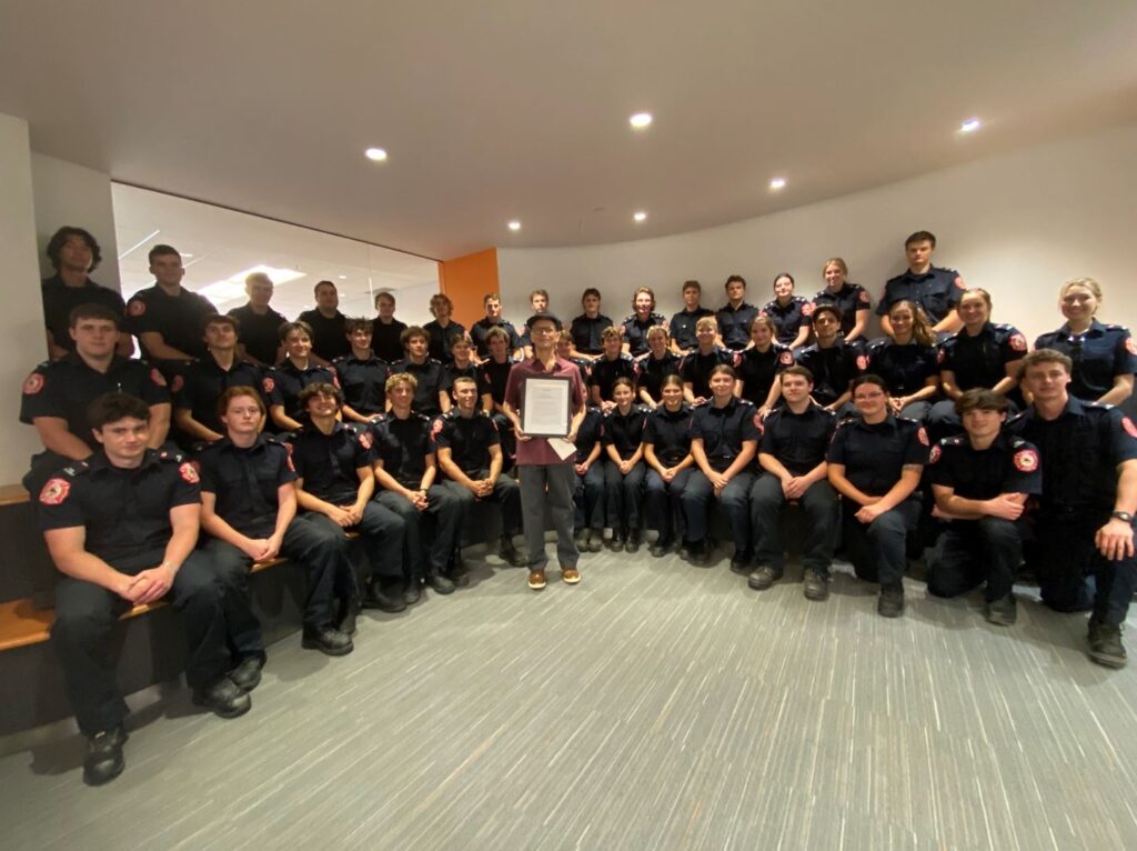 A group of people wearing black uniforms sit on three rows of benches, with one person standing in the centre holding a framed certificate.