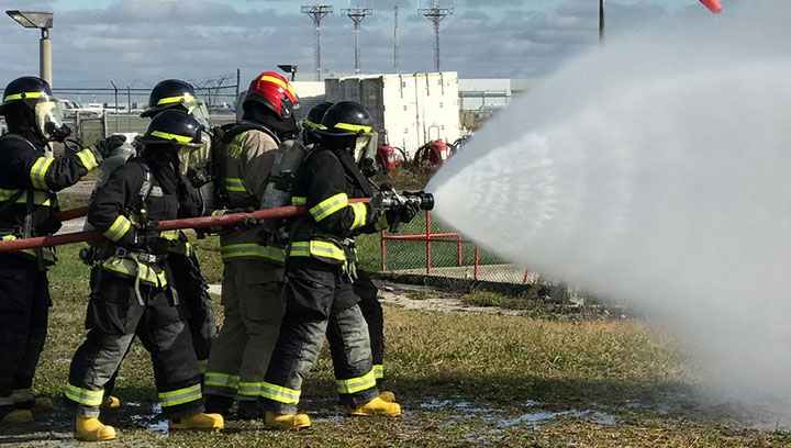 A group of Georgian College firefighting students in uniform holding a hose and spraying water on a fire outdoors