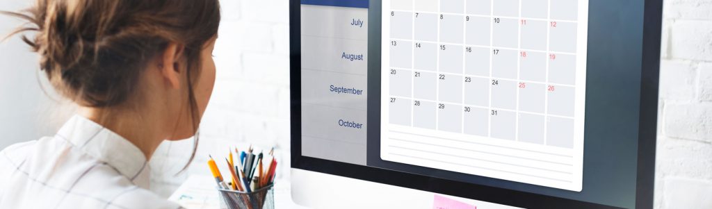 Project manager in a collared shirt and pinned-back hair sitting at a wooden desk while looking at a calendar on a computer