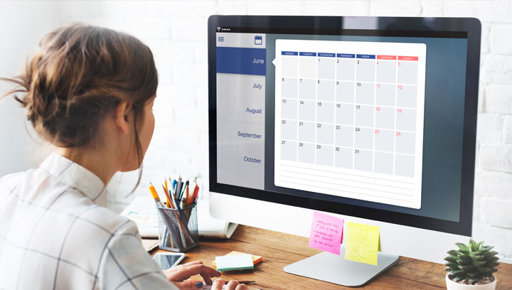 Project manager in a collared shirt and pinned-back hair sitting at a wooden desk while looking at a calendar on a computer