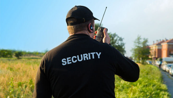 A security officer wearing a uniform jacket, cap and audio earpiece while holding and talking into a handheld transceiver