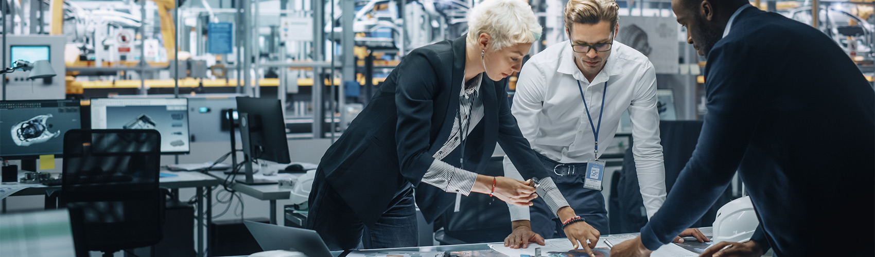 Three people in business attire hunched over a table in a manufacturing plant