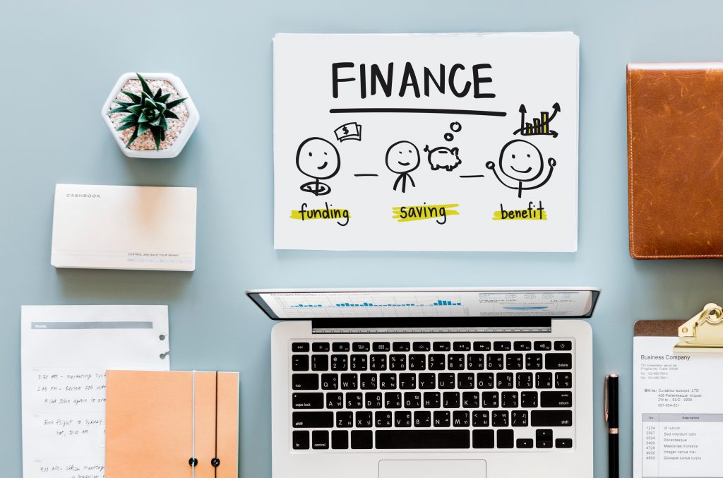Laptop computer on a desk with supplies and a sheet that says finance with images to represent funding, saving and benefit