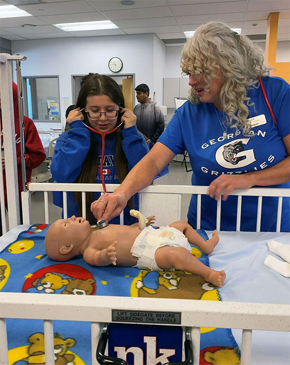 A young female (Audrey Francioni) wearing a stethoscope listening to the heartbeat of an infant simulator in a crib while a blonde older female talks to her.
